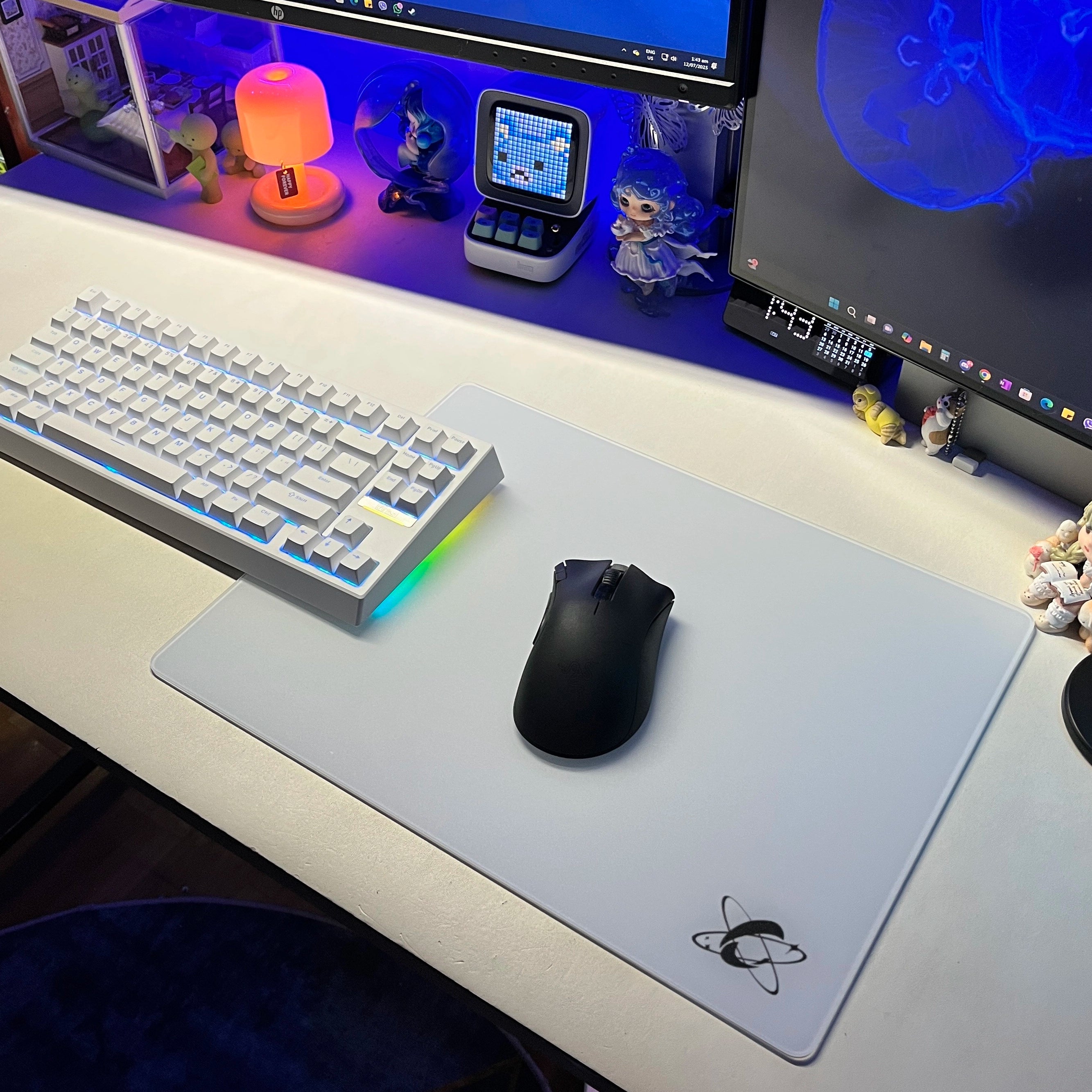 RGB-lit white desk setup featuring the Clustrs Atom White Glass Pad Pro with a black wireless mouse, white mechanical keyboard, and colorful ambient lighting.